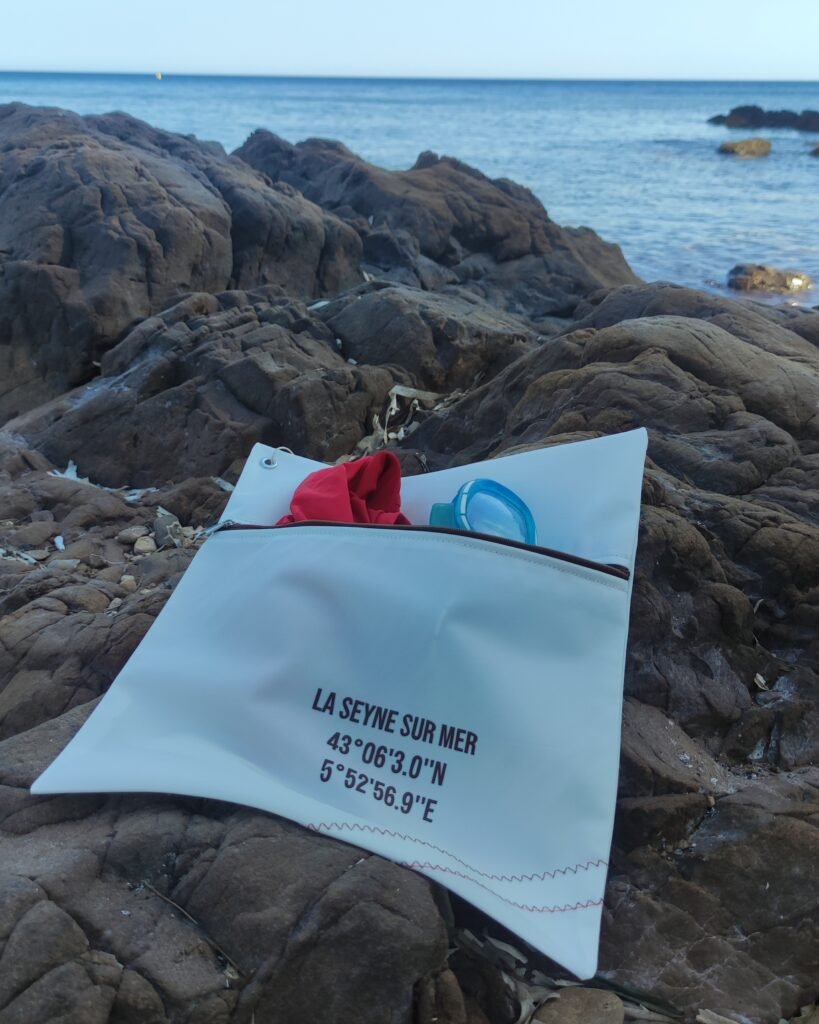 Pochette en voile de bateau recyclée avec fermeture marron et coordonnées de La Seyne-sur-Mer, utilisée pour ranger un maillot de bain et des lunettes à la plage.